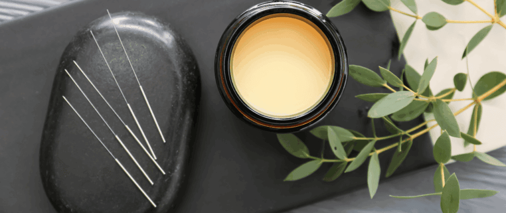 Ein dunkler Stein mit mehreren Nadeln, daneben ein Glas mit ätherischem Öl und grüne Zweige, ideal für Wellness und Aromatherapie.