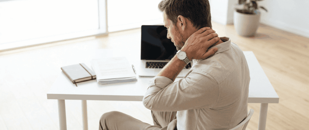 Mann sitzt im Büro und hält sich den Nacken, zeigt Nacken- oder Schulterschmerzen, während er an einem Schreibtisch arbeitet.