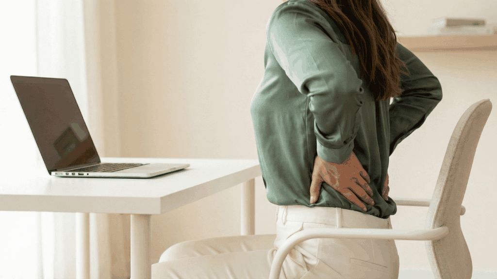 Frau hält sich schmerzhaft den unteren Rücken, sitzt an einem Schreibtisch mit Laptop, zeigt Rückenbeschwerden.