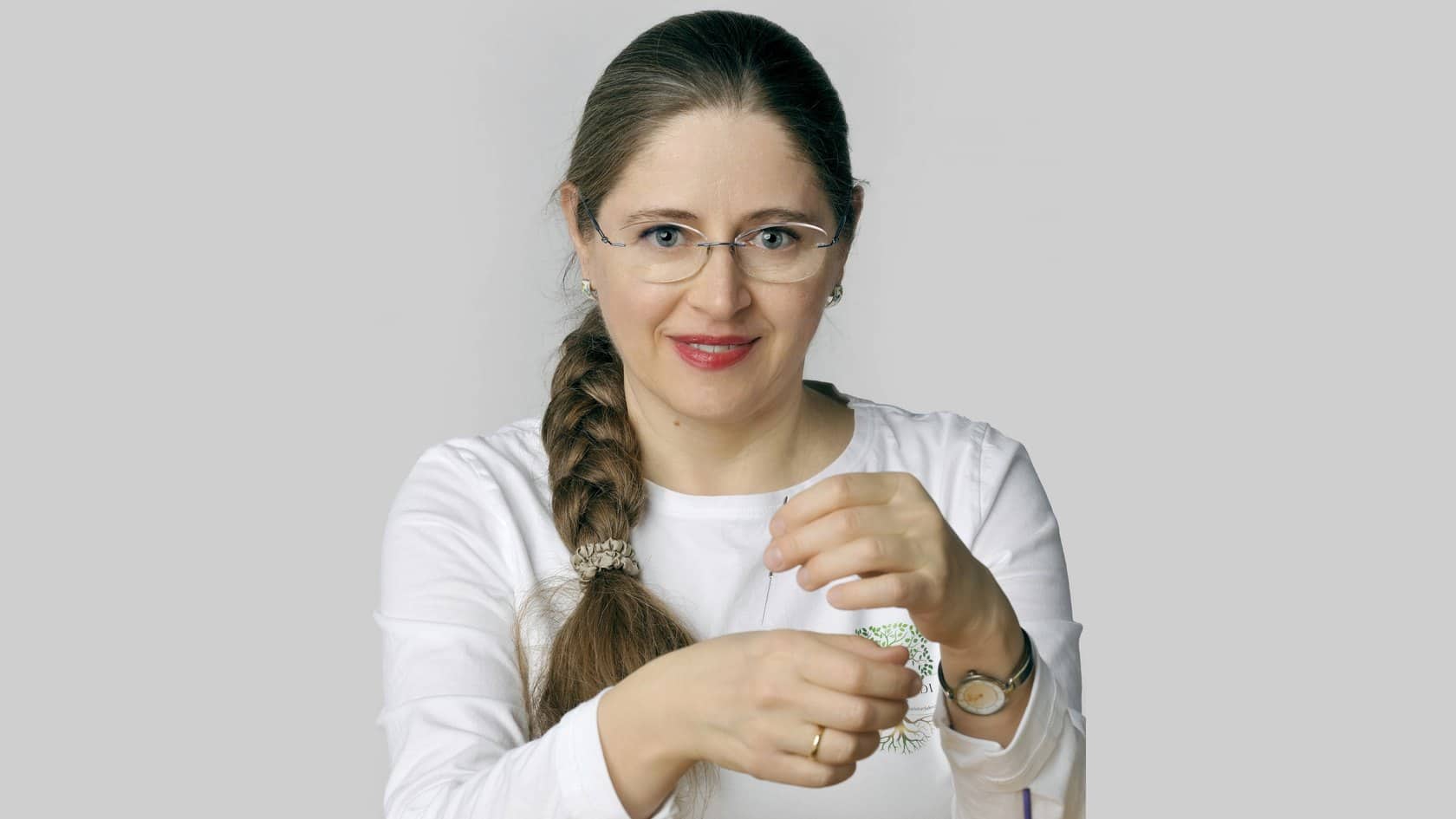 Frau mit Brille und braunem Zopf, lächelnd, in weißem Hemd, vor hellem Hintergrund.